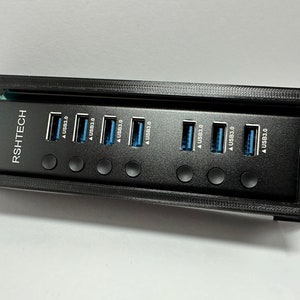 Könnte beinhalten: Schwarzer USB 3.0-Hub mit sieben Anschlüssen und dem Text "RSHTECH". Jeder Anschluss hat ein blaues Innenleben. Der Hub ist in einem schwarzen Rahmen mit Befestigungslöchern an jeder Seite montiert. Die Anschlüsse sind mit "USB3.0" beschriftet.