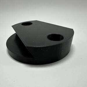 Könnte beinhalten: Ein schwarzes, 3D-gedrucktes Objekt aus Kunststoff mit zwei Löchern und einer ebenen Fläche. Das Objekt ist so konzipiert, dass es an einer Wand oder einer anderen Oberfläche befestigt werden kann.