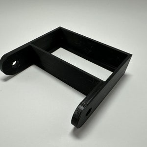 Könnte beinhalten: Schwarzes, 3D-gedrucktes Kunststoffhalterung mit zwei Löchern. Die Halterung hat eine rechteckige Öffnung in der Mitte.