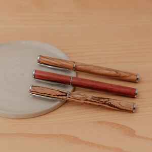 Könnte beinhalten: Drei Holzstift mit silbernen Akzenten auf einer hellbraunen Oberfläche. Die Stifte sind aus verschiedenen Holzarten gefertigt, mit unterschiedlichen Maserungen und Farben.