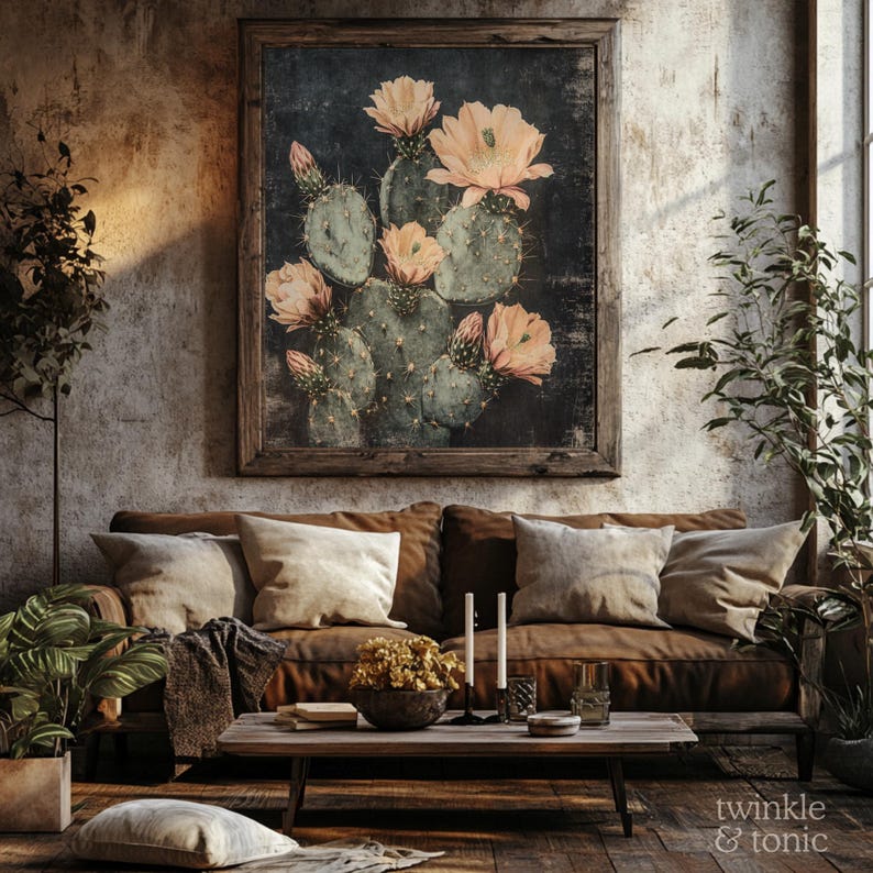 May include: A framed botanical print featuring a cactus with peach-colored flowers. The artwork hangs above a brown leather sofa with beige pillows. The room has a rustic, bohemian aesthetic with wooden furniture and plants.