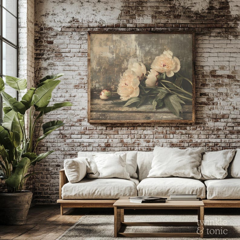 May include: A framed floral print featuring soft, creamy peonies with green foliage. The artwork hangs on a weathered brick wall above a light-colored sofa with white pillows. A wooden coffee table and a potted plant complete the cozy interior.