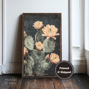 May include: A framed botanical print featuring a cactus with green pads and peach-colored flowers. The artwork has a dark background and a distressed, vintage aesthetic. The print is displayed against a white wall and wooden floor.