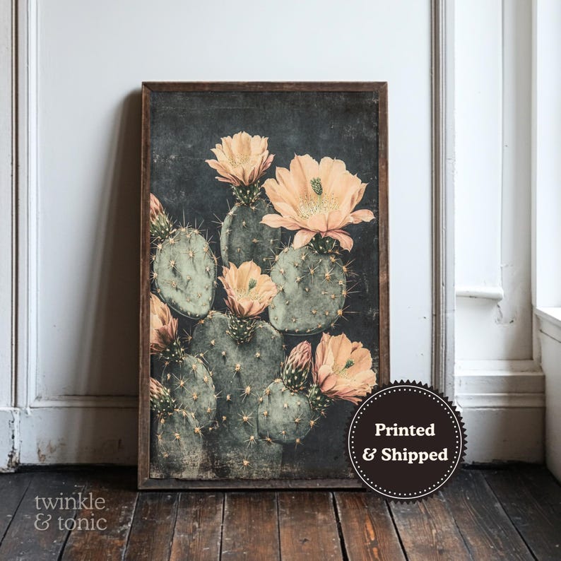 May include: A framed botanical print featuring a cactus with green pads and peach-colored flowers. The artwork has a dark background and a distressed, vintage aesthetic. The print is displayed against a white wall and wooden floor.
