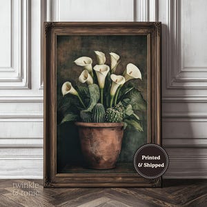 Könnte beinhalten: Gerahmter Kunstdruck mit weißen Calla-Lilien und Kakteen in einem Terrakotta-Topf. Das Kunstwerk hat eine Vintage-Ästhetik mit dunklem Hintergrund und einem braunen Holzrahmen. Der Druck ist mit "twinkle & tonic" beschriftet.