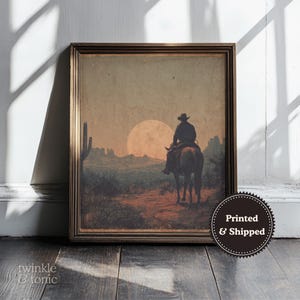 May include: Framed vintage print of a cowboy on horseback silhouetted against a sunset in a desert landscape. The artwork features a warm color palette with a large sun and a cactus. The frame is brown. The print is labeled "Printed & Shipped".