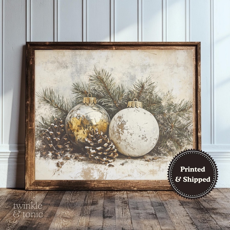 Op de afbeelding: Een ingelijste print met twee kerstballen, &eacute;&eacute;n goudkleurig en &eacute;&eacute;n wit, met dennennaalden en dennenappels. Het kunstwerk heeft een vintage esthetiek, met een bruine houten lijst. De tekst "Printed & Shipped" staat rechtsonder.