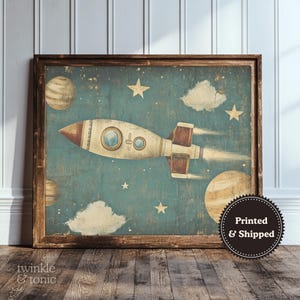 May include: A framed vintage-style print featuring a rocket ship soaring through a starry sky with planets and clouds. The rocket is cream and brown, set against a teal background. The print has a distressed look.