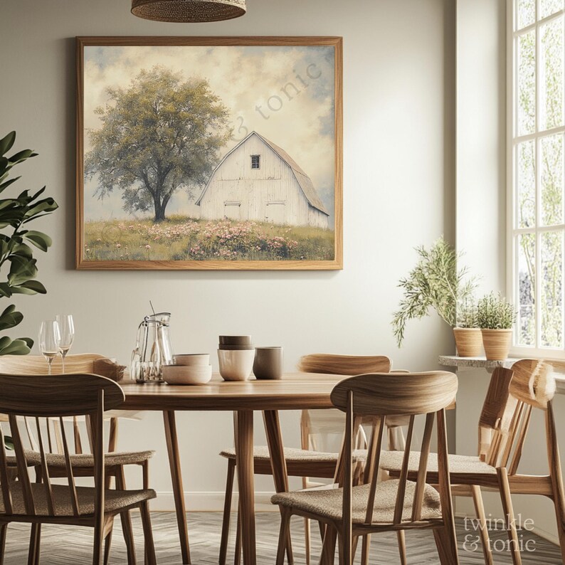 Op de afbeelding: Een ingelijste landschapsafdruk toont een witte schuur en een grote boom in een bloemenveld. Het kunstwerk heeft een houten lijst en hangt boven een houten eettafel met stoelen. De kamer heeft een neutraal kleurenpalet met natuurlijk licht.