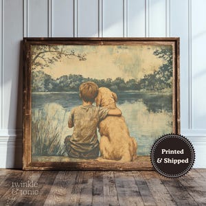 Op de afbeelding: Een ingelijste prent toont een jongen met een golden retriever aan een meer. Het kunstwerk heeft een vintage esthetiek met zachte kleuren en een sereen landschap op de achtergrond. De prent zit in een bruine houten lijst.