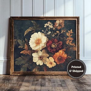 Op de afbeelding: Een ingelijste bloemenprint met een boeket bloemen in crème, bordeauxrood en oranje tegen een donkere achtergrond. Het kunstwerk zit in een houten lijst en wordt op een houten vloer getoond.