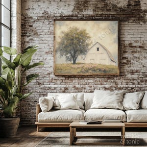 Op de afbeelding: Een ingelijst landschapsschilderij toont een witte schuur naast een grote boom, tegen een zachte, bewolkte lucht. Het kunstwerk staat in een rustieke houten lijst en hangt aan een bakstenen muur boven een lichtgekleurde bank. De sc&egrave;ne roept landelijke rust op.