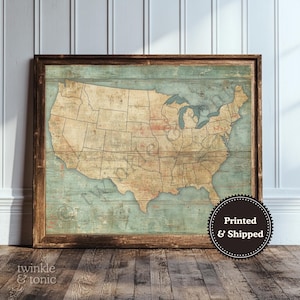 May include: A framed vintage-style map of the United States. The map is in shades of tan, brown, and blue, with state outlines visible. The frame is a dark wood. The map is leaning against a white wall and wooden floor.