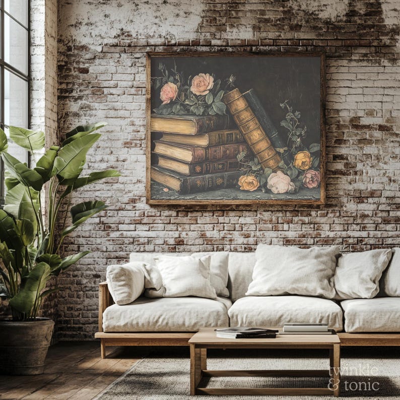 Puede incluir: Una pintura enmarcada representa una naturaleza muerta de libros antiguos y rosas. Los libros est&aacute;n apilados, con cubiertas envejecidas y p&aacute;ginas doradas. Rosas rosas y amarillas est&aacute;n esparcidas alrededor de los libros. La pintura cuelga de una pared de ladrillo sobre un sof&aacute; color crema.