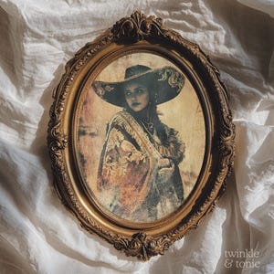 May include: An oval, antique-style gold frame surrounds a vintage print of a person wearing a large hat and shawl. The print has a sepia tone, and the frame features ornate detailing. The text "twinkle & tonic" is in the bottom right corner.