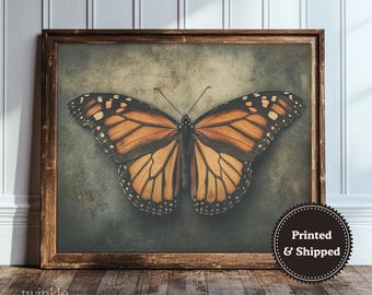 May include: A framed wall art print featuring a monarch butterfly with orange and black wings centered on a dark, textured, olive-green background. The wooden frame has a rustic brown finish. A circular badge in the corner reads Printed & Shipped.