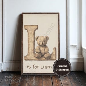 Könnte beinhalten: Gerahmter Kunstdruck für das Kinderzimmer mit einem Teddybären auf dem Buchstaben "L". Der Druck hat eine Vintage-Ästhetik mit dem Text "L is for Liam". Das Kunstwerk befindet sich in einem Holzrahmen.