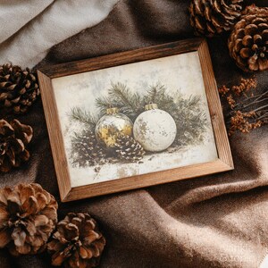 Op de afbeelding: Een ingelijste prent met twee kerstballen en dennenappels. E&eacute;n bal is goudkleurig, de andere is wit. De prent zit in een houten lijst, geplaatst op een bruin stoffen oppervlak, met dennenappels verspreid rond de lijst.