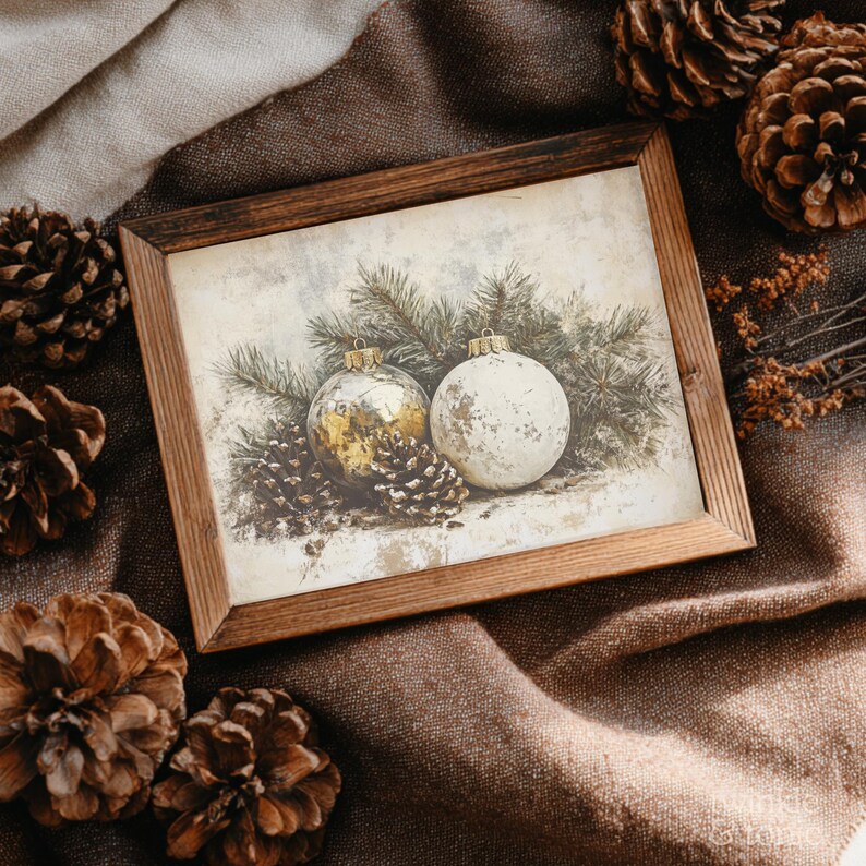 Op de afbeelding: Een ingelijste prent met twee kerstballen en dennenappels. E&eacute;n bal is goudkleurig, de andere is wit. De prent zit in een houten lijst, geplaatst op een bruin stoffen oppervlak, met dennenappels verspreid rond de lijst.