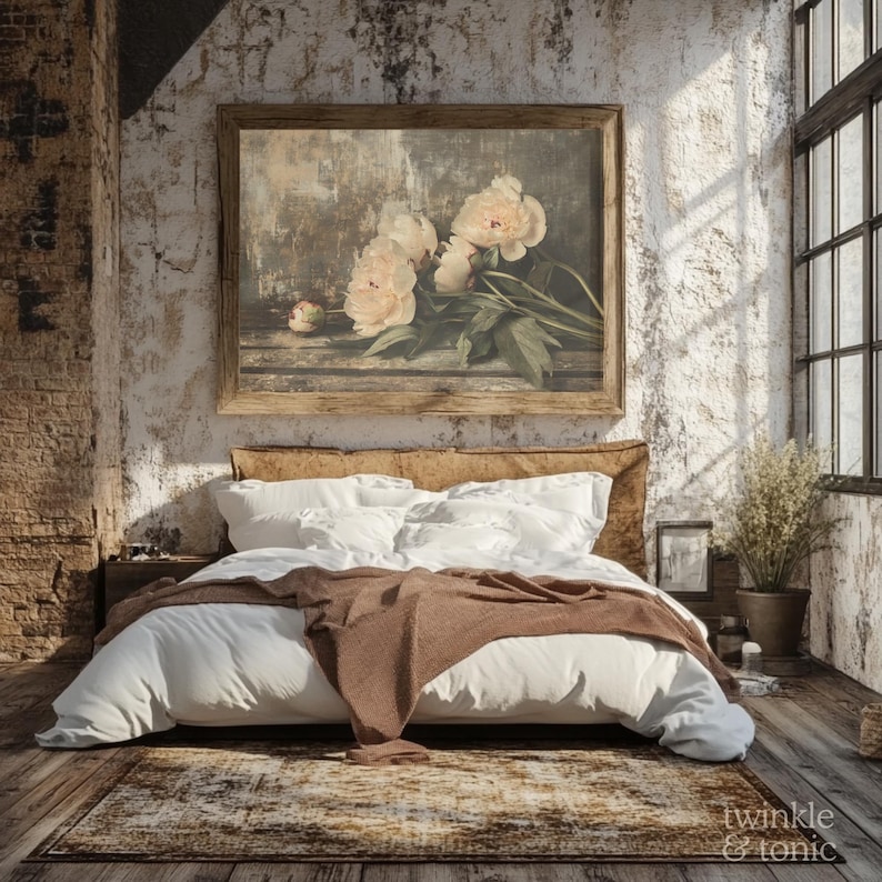 May include: A bedroom scene featuring a bed with white bedding and a brown throw blanket. Above the bed is a framed floral print with cream-colored peonies. The room has a rustic, vintage aesthetic with exposed brick and a large window.