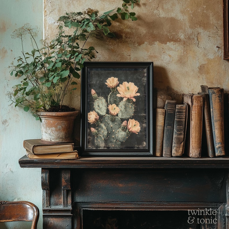 May include: A framed print of a cactus with peach-colored flowers. The artwork is displayed on a dark wooden mantelpiece, alongside antique books and a potted plant. The image has a vintage aesthetic.