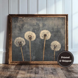 May include: A framed print featuring four dandelion seed heads against a muted blue-gray background. The artwork has a vintage aesthetic, with a distressed look. The frame is a warm brown color. The print is displayed on a wooden floor.
