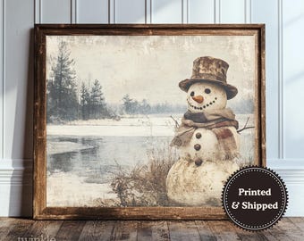 May include: A framed print of a snowman in a winter landscape. The snowman wears a top hat, scarf, and has a carrot nose. The background shows a snowy scene with trees and a frozen lake. The frame is made of wood.