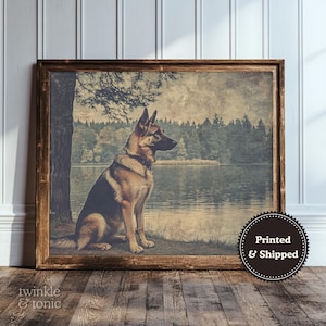 May include: A framed print featuring a German Shepherd dog sitting by a lake. The dog is brown and black, with a focused gaze. The artwork has a vintage, faded aesthetic, with a tree and forest in the background. The frame is made of dark wood.