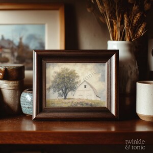 Op de afbeelding: Een ingelijste prent met een witte schuur en een grote boom op een grasheuvel onder een bewolkte hemel. De lijst is donkerbruin. De prent wordt tentoongesteld op een houten oppervlak met andere decoratieve items.