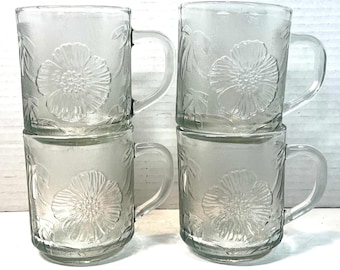 May include: Four clear glass mugs stacked in two pairs. Each mug features a floral design with a handle. The floral pattern is embossed on the glass, creating a textured effect. The mugs are suitable for serving hot or cold beverages.