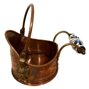 May include: A copper-colored metal coal scuttle with a curved handle and a decorative handle with a white and blue floral design. The scuttle has a small, decorative brass accent on the side.
