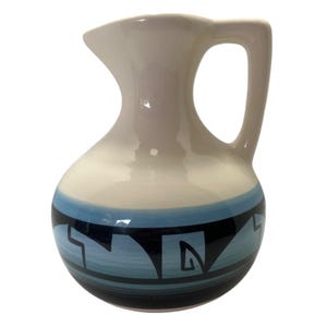 May include: A small, ceramic pitcher with a curved spout and handle. The pitcher has a white top that fades into a light blue band with a black geometric design. The bottom of the pitcher is a darker blue. The pitcher is likely used for serving liquids.