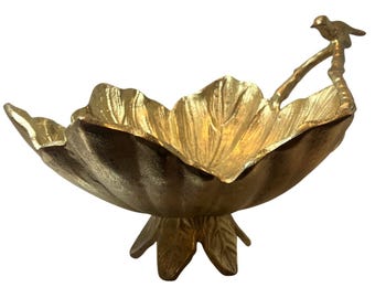 May include: A gold-colored metal bowl shaped like a large, open flower petal with a textured surface. The bowl features a small bird perched on a branch-like handle extending from one side and rests on a base shaped like smaller leaves.