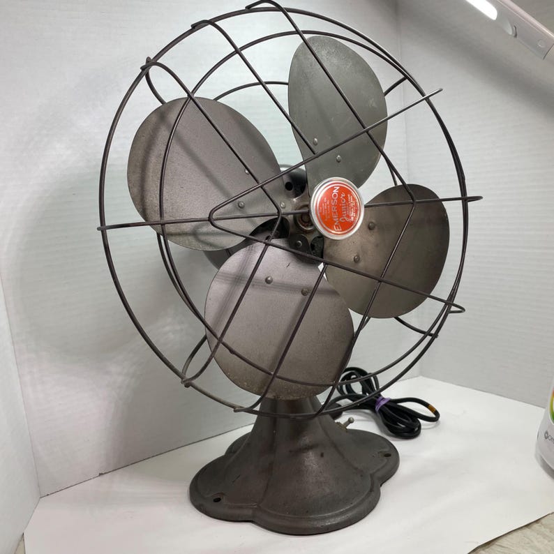 May include: An antique Emerson Junior oscillating fan. The fan has three blades and is encased in a wire cage. The base is a dark, ornate metal. The fan has a red and orange emblem in the center.