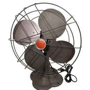 May include: An antique electric fan with a dark gray finish. The fan has four blades, a protective wire cage, and a red center piece. The base is a decorative, rounded shape. The power cord is black.