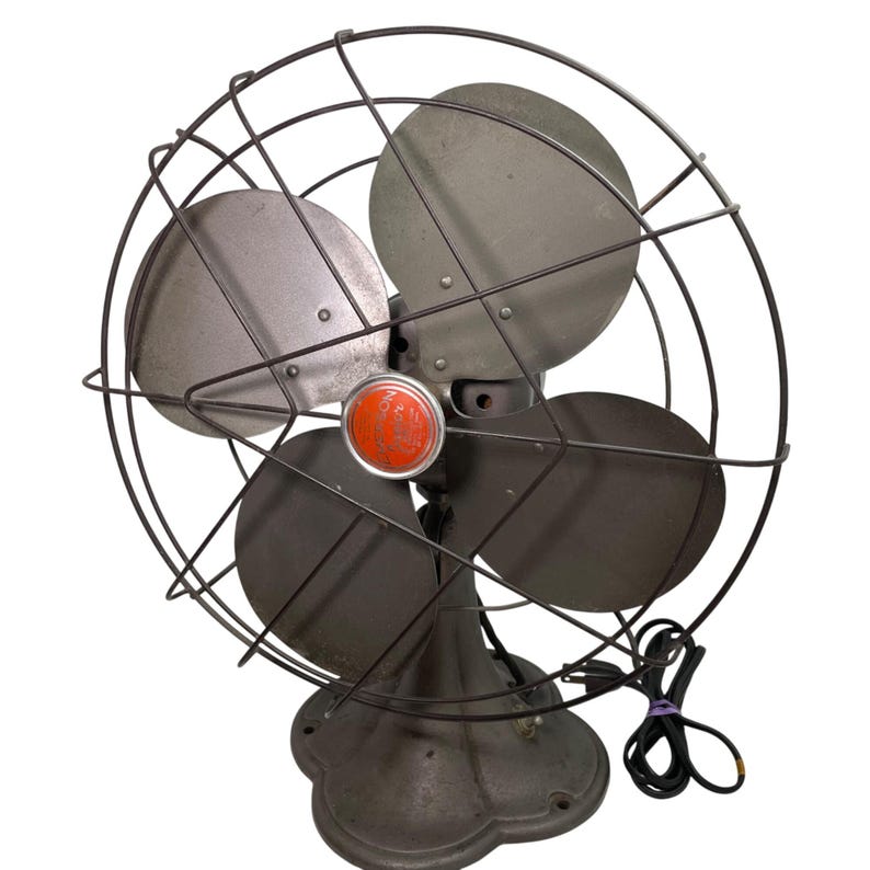 May include: An antique electric fan with a dark gray finish. The fan has four blades, a protective wire cage, and a red center piece. The base is a decorative, rounded shape. The power cord is black.