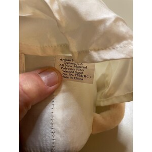 May include: White stuffed doll with a white fabric tag that reads "Artisan F Oxnard, CA All New Material Polyester Fiber Washed Sand No. PA-7798(RC) Made in China"
