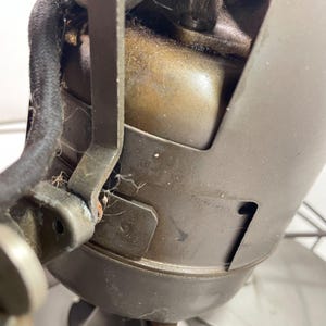 May include: Close-up of an antique electric fan motor. The motor housing is a dark, aged metal with visible wear and tear. A black, cloth-covered power cord is attached. The fan blades are partially visible.