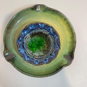 May include: A ceramic ashtray with a layered design. The outer layer is a green-yellow hue, the middle layer is blue and brown, and the center has a green, textured appearance. The ashtray has a curved shape with a small rest.