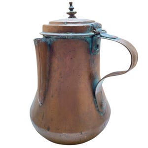 May include: A vintage copper pitcher with a handle and a lid. The pitcher has a bulbous body and a spout. The copper has a patina with areas of green and blue oxidation. The handle is curved and attached to the side of the pitcher. The lid has a decorative finial.