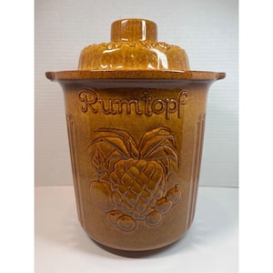 May include: A brown ceramic Rumtopf pot with a lid. The pot features embossed fruit designs and the word "Rumtopf" in script. The lid has a small handle and a decorative rim. The pot is designed for preserving fruit in rum.