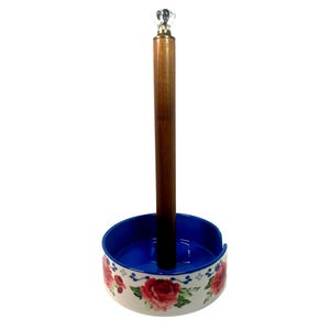 May include: A paper towel holder with a wooden center post and a decorative base. The base is white with a blue interior and a floral pattern featuring red roses and green leaves. The top of the post has a clear, faceted crystal-like finial.