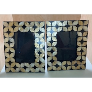 May include: Two black picture frames with a decorative pattern of gold and cream-colored shell-like shapes. The frames have a square shape with a black interior space for photos. The frames are placed side by side.