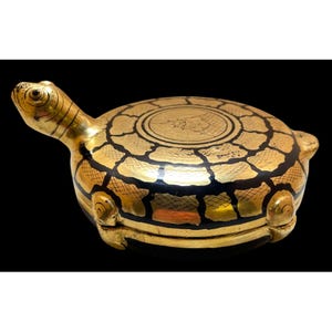 May include: A decorative turtle-shaped trinket box. The box is primarily gold with black accents, featuring a detailed shell pattern and a turtle head extending from the front. The box is on a black background.