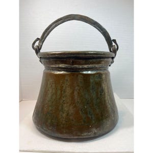 May include: A bronze-colored metal cauldron with a curved handle. The cauldron has a wide base that tapers towards the top. The handle is attached to the rim with metal loops. The metal has a mottled appearance with shades of brown and green.