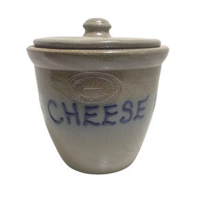 May include: A vintage stoneware cheese crock with a lid. The crock is a light gray color with blue lettering that spells out "CHEESE". The crock has a round shape and a small knob on the lid. The words "Lexington Pottery York, Maine" are stamped on the crock.