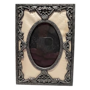 May include: An ornate, rectangular picture frame with a silver-colored, detailed border. The frame has an oval opening in the center and a cream-colored background. The frame is designed with floral and scrollwork patterns.