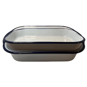 May include: Two rectangular enamel baking pans stacked on top of each other. Each pan features a smooth white interior and exterior surface with a contrasting dark blue rim around the top edge. The pans have rounded corners and a shallow depth.