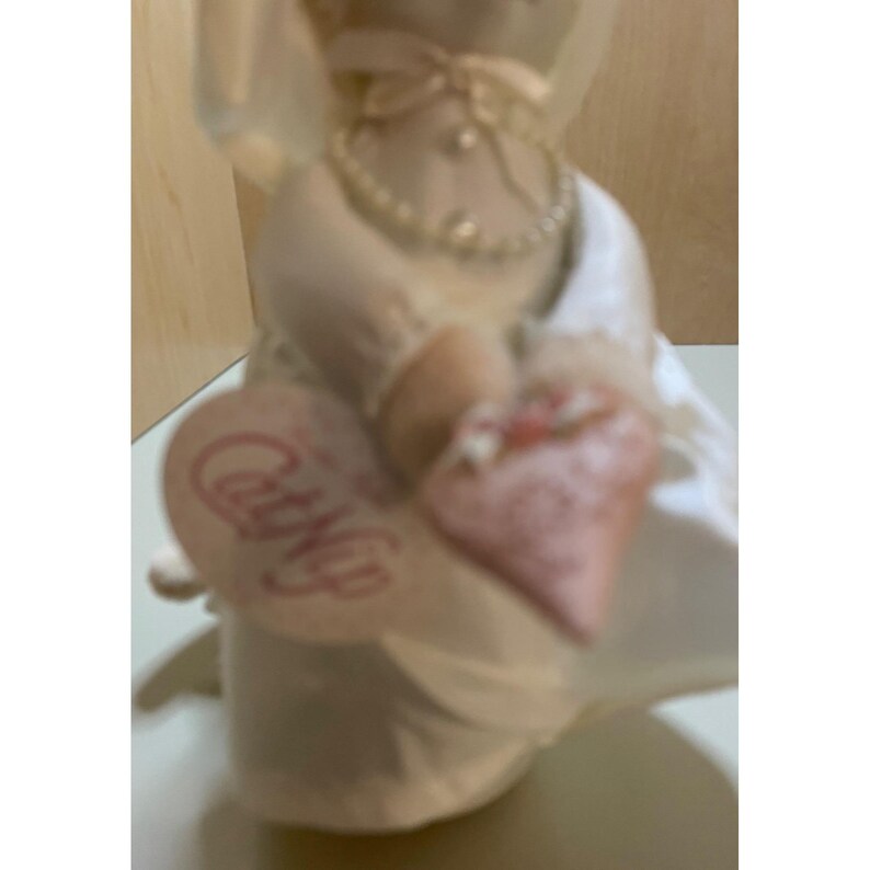 May include: A white porcelain figurine of a bride wearing a white dress and veil, holding a small pink heart-shaped pillow with the word "Carry Me" printed on it.