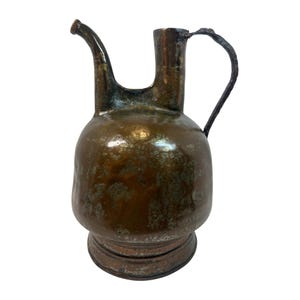 May include: A vintage copper pitcher with a spout and handle. The pitcher has a bulbous body and a dark, aged patina. The handle is a simple, curved design. The spout is curved and extends from the top of the pitcher.